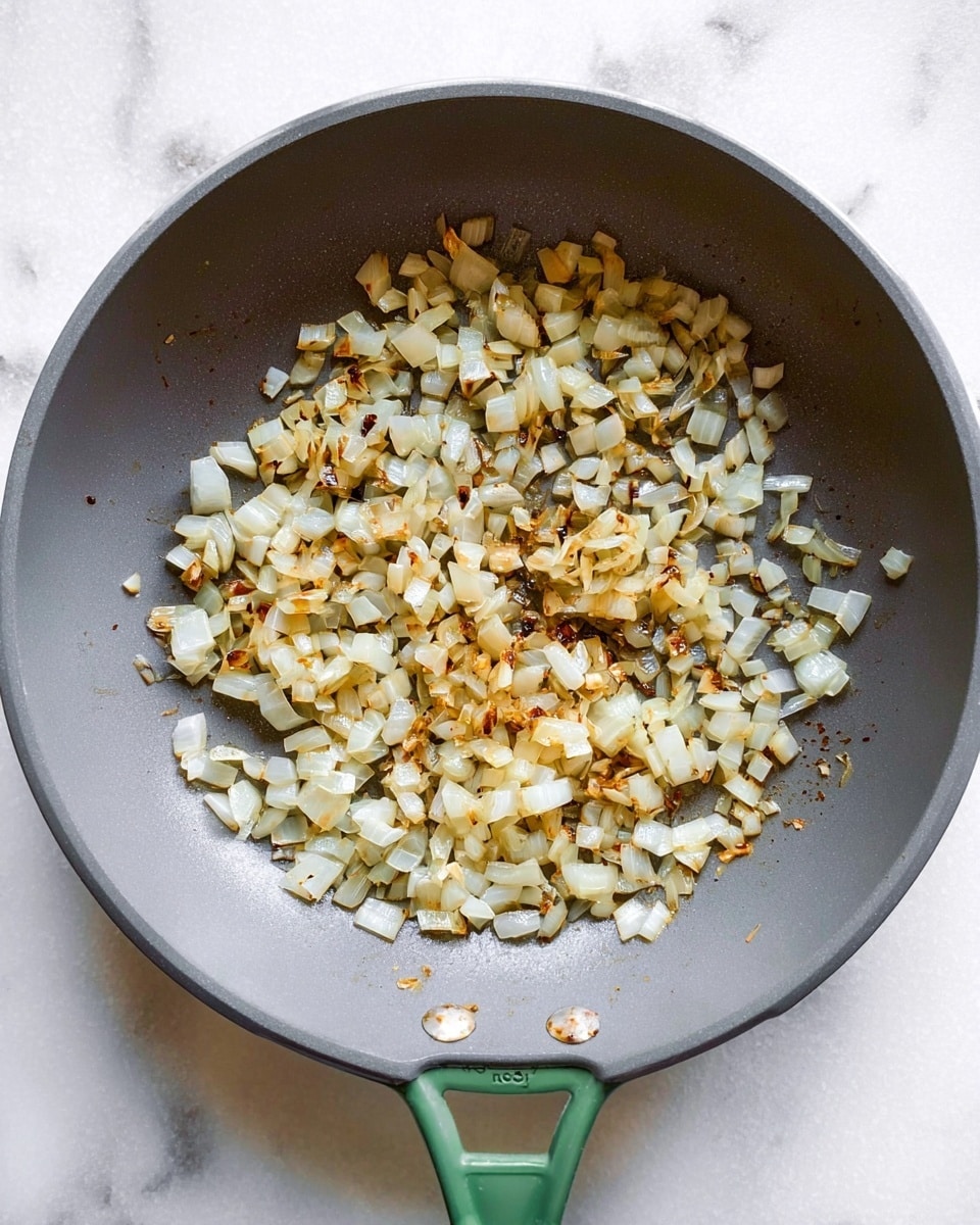 A top view of a shallow gray pan with a green handle on a white marbled surface, filled with one layer of small, diced sautéed onions that are light golden brown with some darker caramelized spots, spread mostly in the center and right side of the pan, showing their soft texture and slight shine from cooking oil photo taken with an iphone --ar 4:5 --v 7