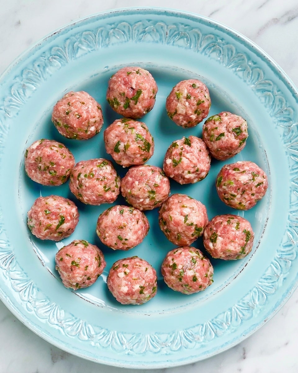 The image shows a light blue large round plate with a raised decorative design around the edge, covered with fifteen raw meatballs arranged in a semi-random pattern. Each meatball is pink with visible small green herb pieces mixed evenly throughout. The meatballs have a smooth texture and are about the same size, about the size of small golf balls. The plate sits on a white marbled surface. photo taken with an iphone --ar 4:5 --v 7