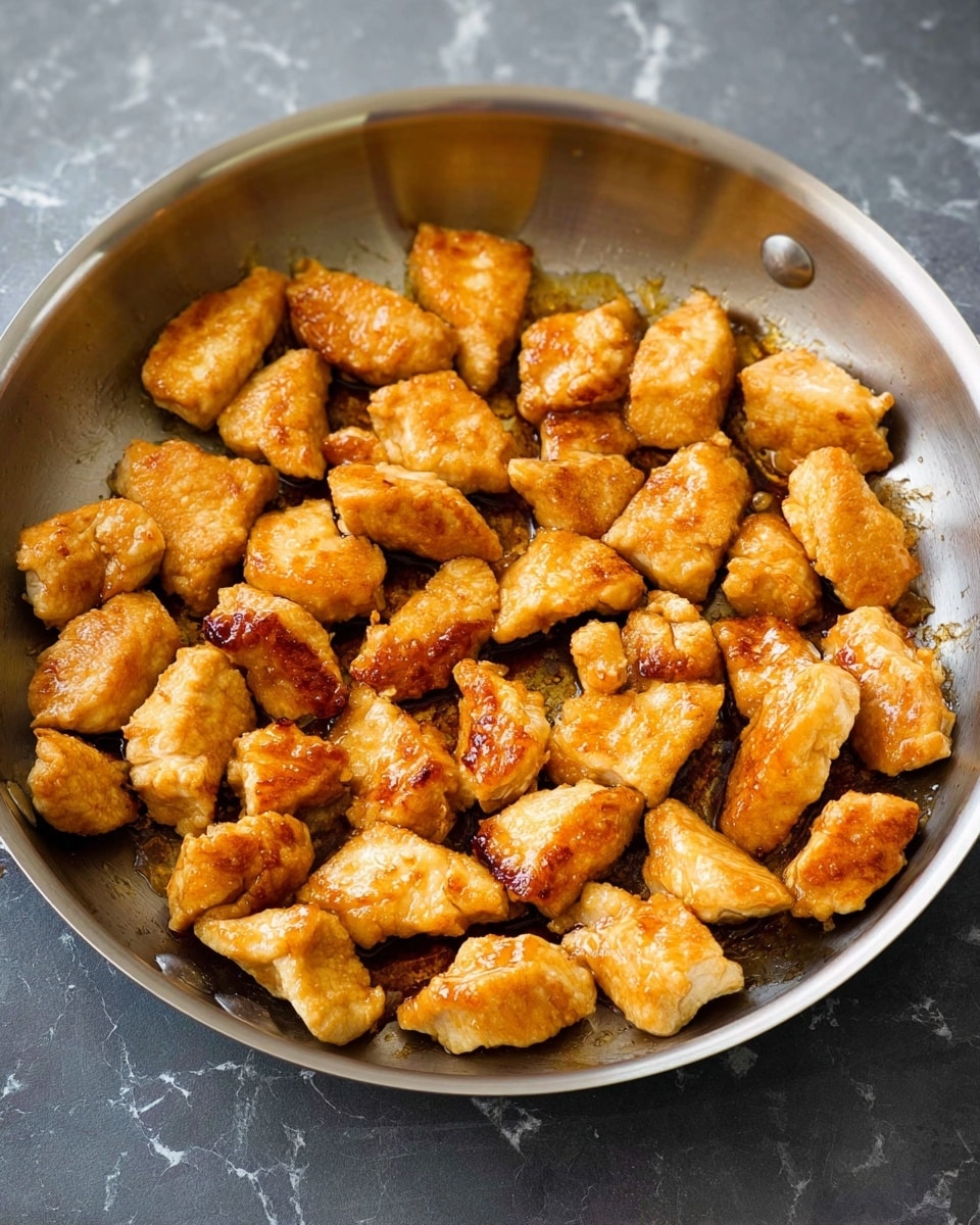 A silver pan filled with about 30 small, thick pieces of cooked chicken, each piece golden brown with a slight shine indicating cooked juices or light sauce. The chicken pieces are unevenly shaped but roughly similar in size, covering most of the pan's surface with some gaps showing the inside of the pan. The pan sits on a dark, textured surface, but the surrounding background is changed to a white marbled texture. The overall look is simple and appetizing, focusing on the cooked chicken details. Photo taken with an iphone --ar 4:5 --v 7