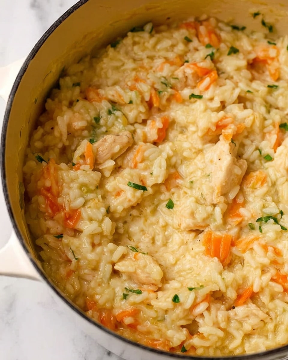 The image shows a close-up of a creamy rice dish inside a white cooking pot. The rice is soft and pale yellow, mixed with small pieces of orange carrot and chunks of light brown chicken. The texture looks slightly creamy and moist, with small bits of green herbs scattered throughout. The pot has a smooth white inside and is set against a white marbled surface in the background. photo taken with an iphone --ar 4:5 --v 7