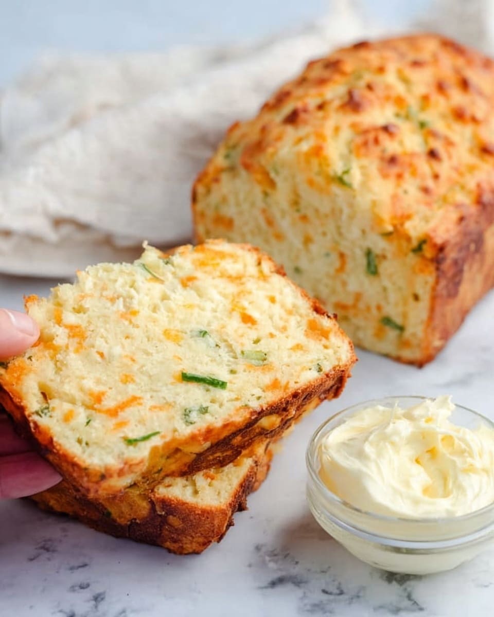 The image shows two thick slices of cheesy bread resting on a white marbled surface, with a larger piece of the bread behind them. The bread has a golden-brown crust with a slightly rough texture and visible bits of green herbs and orange cheese inside. Next to the bread is a small clear glass bowl filled with creamy softened butter. A woman’s hand is holding one of the slices gently. The background includes a soft white cloth adding a cozy feel. photo taken with an iphone --ar 4:5 --v 7