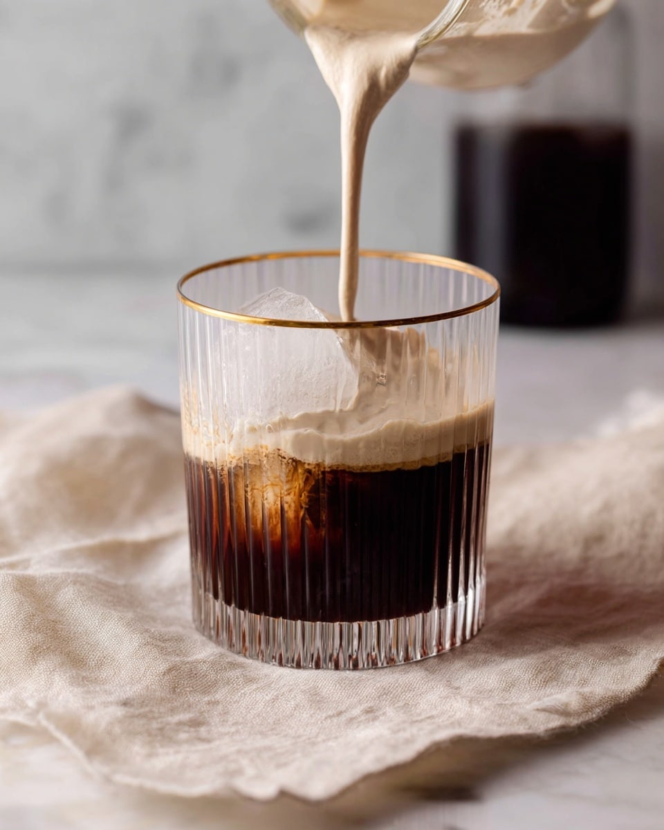A clear glass with vertical ridges and a gold rim is filled halfway with dark cold coffee and a large ice cube inside. A creamy light beige foam layer is being poured on top, slowly covering the coffee with a thick, smooth texture. The glass sits on a soft beige cloth on a white marbled surface. In the background, there is a blurred dark container. Photo taken with an iphone --ar 4:5 --v 7