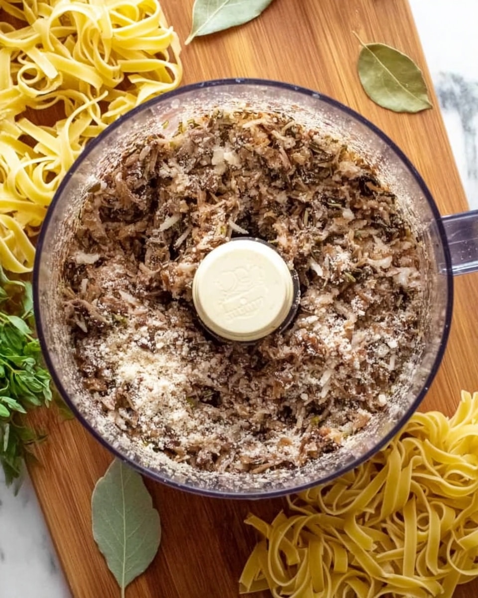 The image shows the top view of a transparent food processor bowl filled with a finely mixed, mostly brown and white chunky mixture. Around the processor, there are uncooked yellow fettuccine pasta nests placed on a wooden cutting board. At the bottom left corner, some green herbs are partially visible, and two light green bay leaves are placed next to the processor. The base surface under the cutting board is a white marbled texture. Photo taken with an iphone --ar 4:5 --v 7