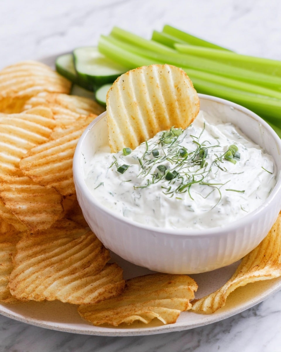 A white bowl filled with thick, creamy white dip topped with small green herb pieces sits in the center. One large ridged potato chip is dipped into the bowl, standing upright. Around the bowl on a white plate, several golden ridged potato chips lie scattered, and behind them are fresh green celery sticks arranged in a small pile. The whole setup is placed on a white marbled surface. photo taken with an iphone --ar 4:5 --v 7