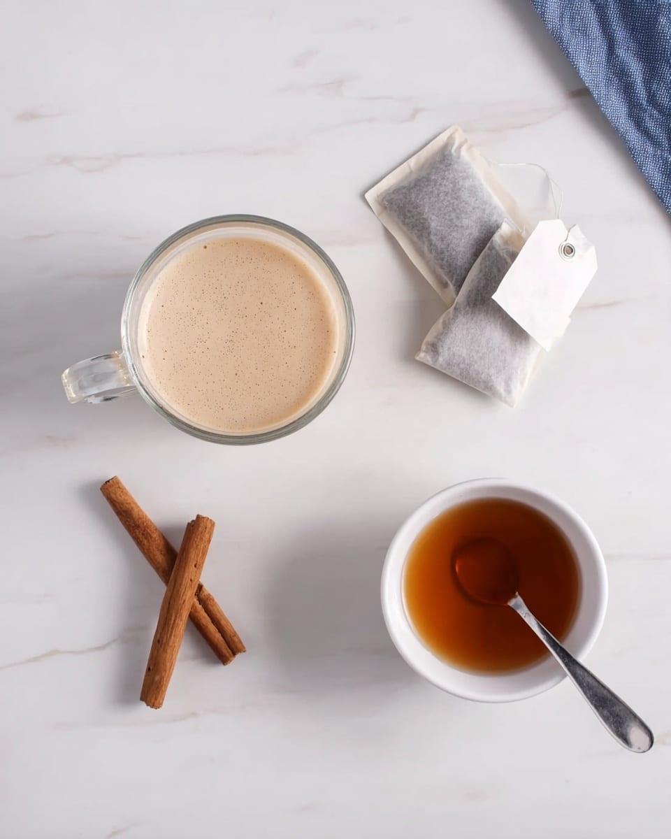 The image shows a clear glass cup filled with a light brown, frothy liquid on a white marbled surface. Next to the cup, there are two tea bags with paper tags, one more beige and one white, lying flat. Two cinnamon sticks are placed side by side near the tea bags. On the right side, there is a small white bowl holding a brown liquid, with a silver spoon resting inside it. The overall setting is clean and simple with soft lighting. photo taken with an iphone --ar 4:5 --v 7