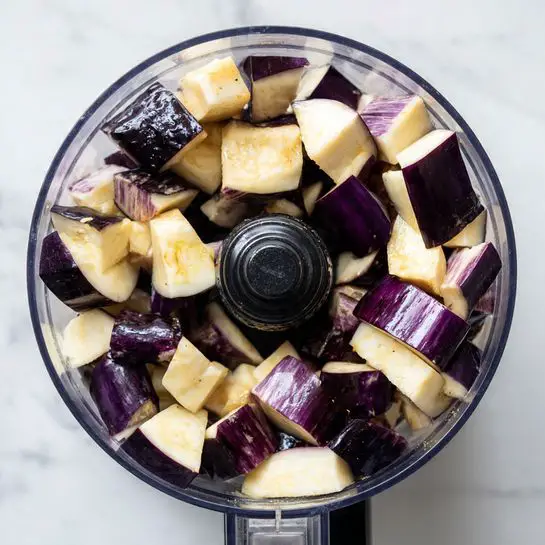 A top view of a clear food processor bowl filled with chopped dark purple and cream pieces of eggplant placed in the center. The bowl rests on a white marbled surface, and the eggplant pieces show a mix of smooth, slightly shiny purple skin and softer, pale inside flesh, unevenly chopped. The black central blade hub is visible under some eggplant pieces. Photo taken with an iphone --ar 4:5 --v 7