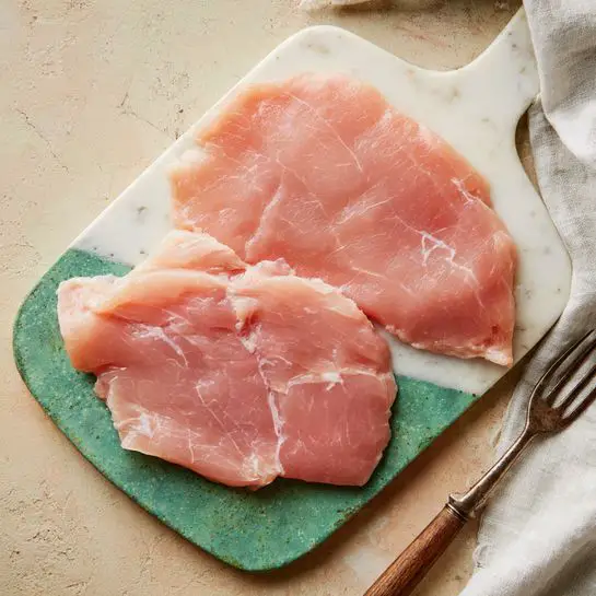 Two large thin pieces of raw chicken with a pale pink color, some light white veins, and a slight shine lay flat on a white cutting board with a green rim. The chicken pieces have an uneven texture with some parts thinner and more translucent than others. The cutting board rests on a beige surface with a rough look. To the right side, a white cloth is casually placed next to an old metal meat fork with a wooden handle. Photo taken with an iphone --ar 4:5 --v 7