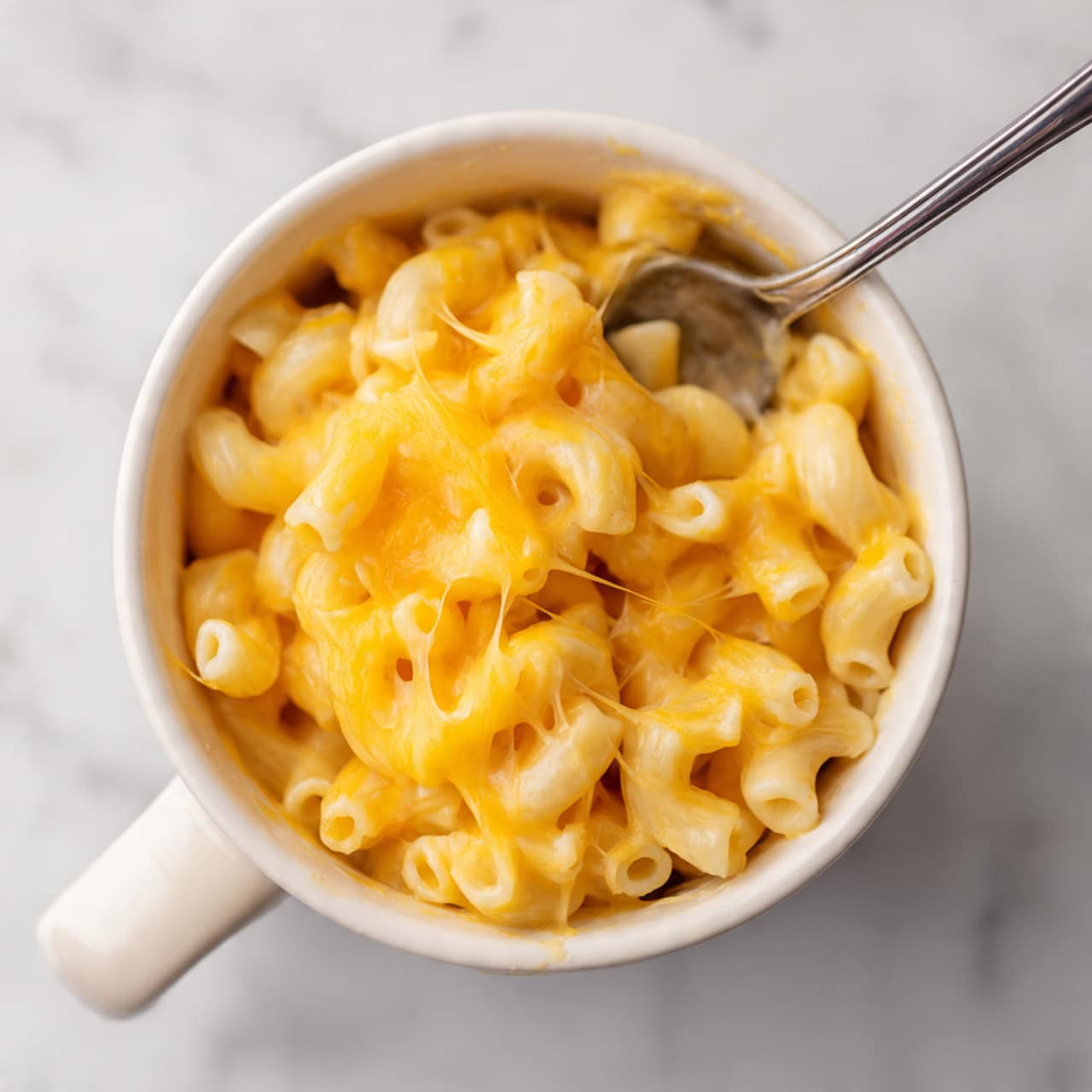 A close-up view of a white mug filled with one layer of bright yellow-orange macaroni and cheese, showing creamy and smooth texture with elbow-shaped pasta evenly coated. Above the mug, a silver fork holds a small scoop of the macaroni, highlighting the cheesy shine and soft pasta curves. The scene is set on a white marbled surface. Photo taken with an iphone --ar 4:5 --v 7