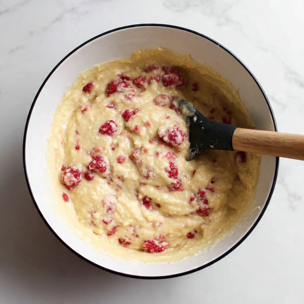 A white bowl with a black rim sits on a white marbled surface, filled halfway with thick, creamy, pale yellow batter that has a smooth texture with small lumps. Red raspberries are mixed into the batter, with some partly visible and others covered. A black spatula with a wooden handle rests inside the bowl, partly covered with the batter. The scene is simple and clean, focused on the mixing process. photo taken with an iphone --ar 4:5 --v 7