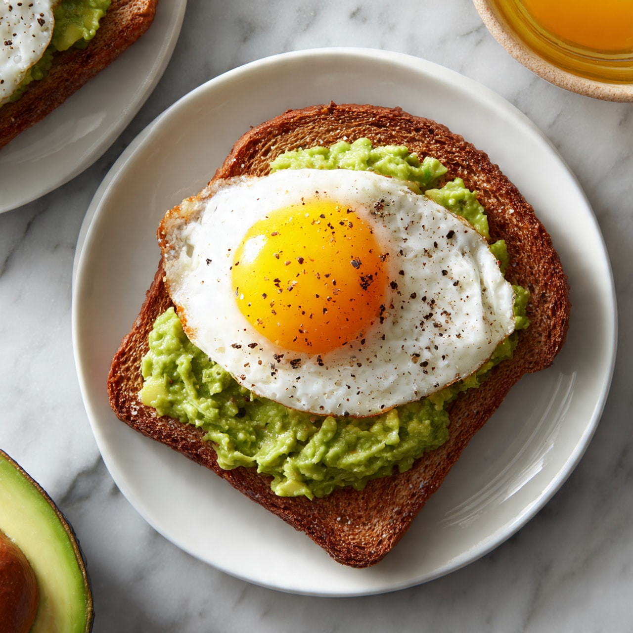The image shows two pieces of brown bread with a textured crust, both topped with a layer of smashed green avocado, giving a fresh and chunky look. On top of the avocado of the closest toast, there is a fried egg with a bright yellow runny yolk spilling slightly over the avocado, and the egg white is cooked but soft, sprinkled with black pepper. Both toasts are placed on a stack of white plates on a white marbled surface. The background is softly blurred, focusing on the front toast. photo taken with an iphone --ar 4:5 --v 7