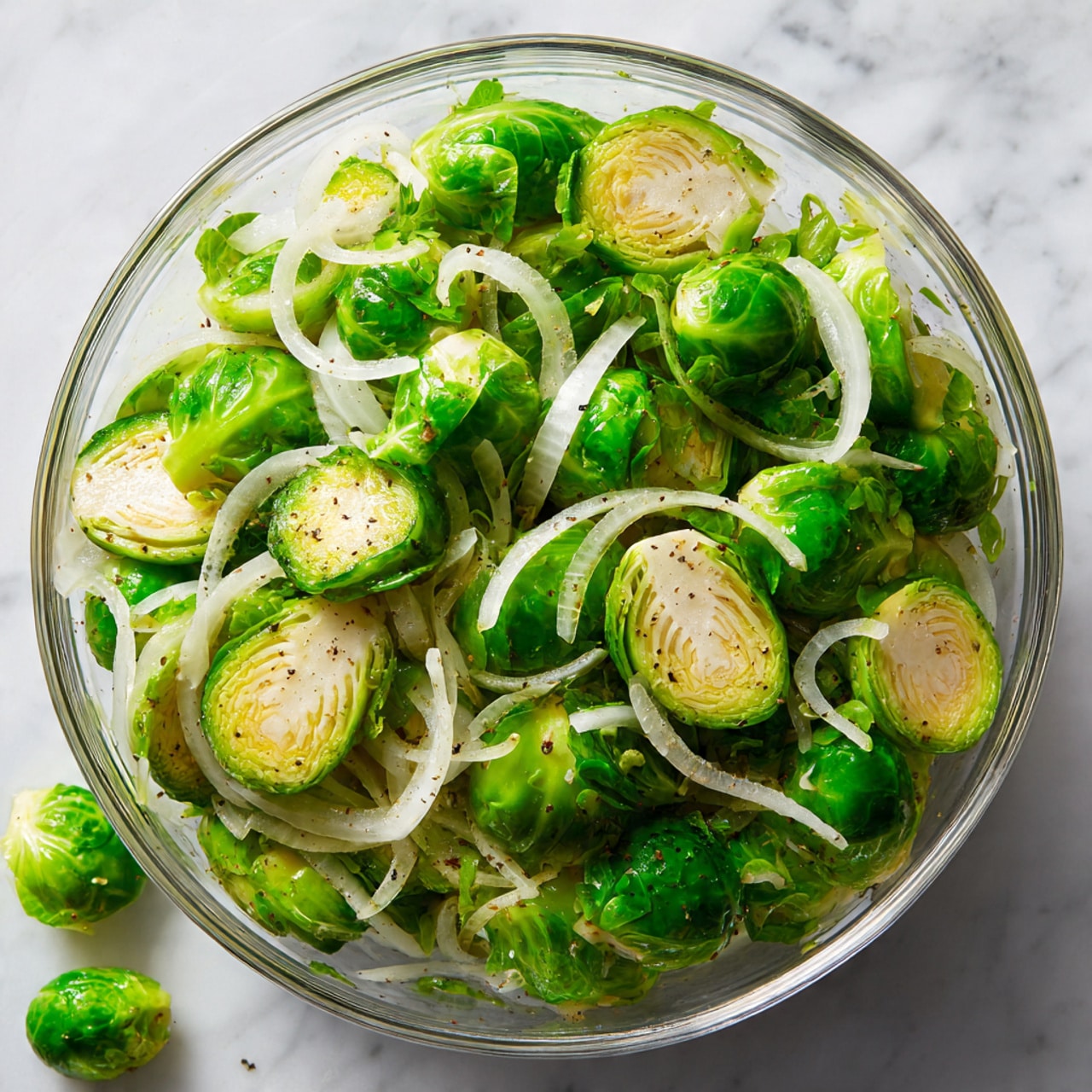 The image shows a clear glass bowl filled with roughly two layers of halved bright green Brussels sprouts and thin white onion slices mixed together. The Brussels sprouts have a fresh and slightly roasted look with visible texture on the cut sides in light beige. The onion slices are curly and sit evenly among the Brussels sprouts, giving a soft white contrast. The bowl is placed on a white marbled surface with a few loose Brussels sprout leaves scattered nearby. The lighting is bright and natural, highlighting the fresh ingredients clearly. Photo taken with an iphone --ar 4:5 --v 7