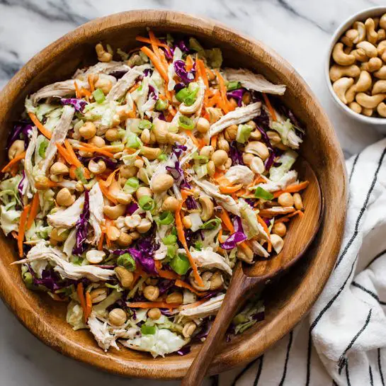 A large wooden bowl filled with a colorful salad showing pieces of cooked chicken, bright orange carrot sticks, chopped purple cabbage, light beige chickpeas, chopped green onions, and cashew nuts scattered on top. A wooden spoon is resting inside the bowl, partially submerged in the salad. The bowl sits on a white marbled surface, with a white cloth with black stripes in the background and a small white bowl filled with cashews to the top right. The textures show the softness of the chicken, crunchiness of the vegetables, and nuttiness of the cashews, all mixed well together photo taken with an iphone --ar 4:5 --v 7
