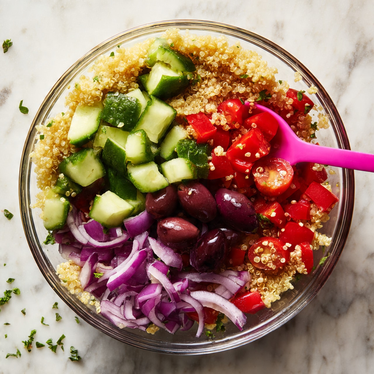 A clear glass bowl holds a colorful salad with layers of chopped quinoa forming the base, topped with bright green cucumber pieces that have a rough outer texture and pale green inside, deep red diced bell peppers, small chunks of vibrant green bell pepper, thin slices of purple onion, halved cherry tomatoes showing bright red interiors, and several dark purple olives scattered on top. A woman's hand pink spoon is partially visible inside the bowl, resting on the right side. The bowl sits on a white marbled surface with a few small green herb bits around. photo taken with an iphone --ar 4:5 --v 7