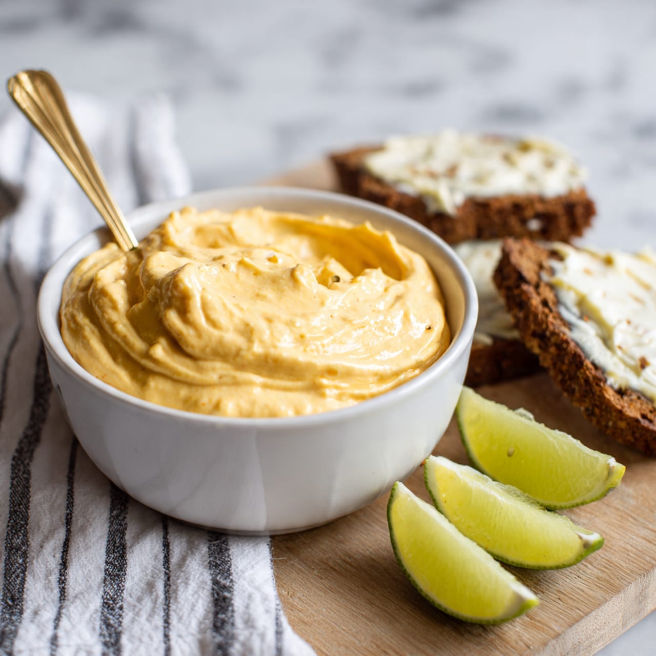 A small white bowl filled with smooth, creamy orange dip that has some small bits inside, showing a soft texture. A golden spoon is placed inside the bowl with some dip on it. Next to the bowl, there are three slices of green lime and pieces of dark brown bread with some white spread on top. The bowl and food are placed on a wooden surface with a white and black striped cloth nearby, all on a white marbled background. Photo taken with an iphone --ar 4:5 --v 7