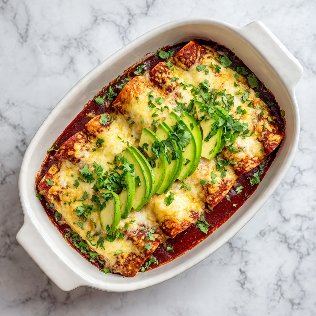 A white plate holds a single enchilada with a golden-brown baked cheese layer on top, slightly melted and textured. The enchilada is covered with a rich reddish-brown sauce that seeps into the edges. On top, there is a fresh slice of bright green avocado and chopped green cilantro leaves scattered across the dish and plate. The plate sits on a white marbled surface with a black and white patterned cloth partly visible nearby. Photo taken with an iphone --ar 4:5 --v 7