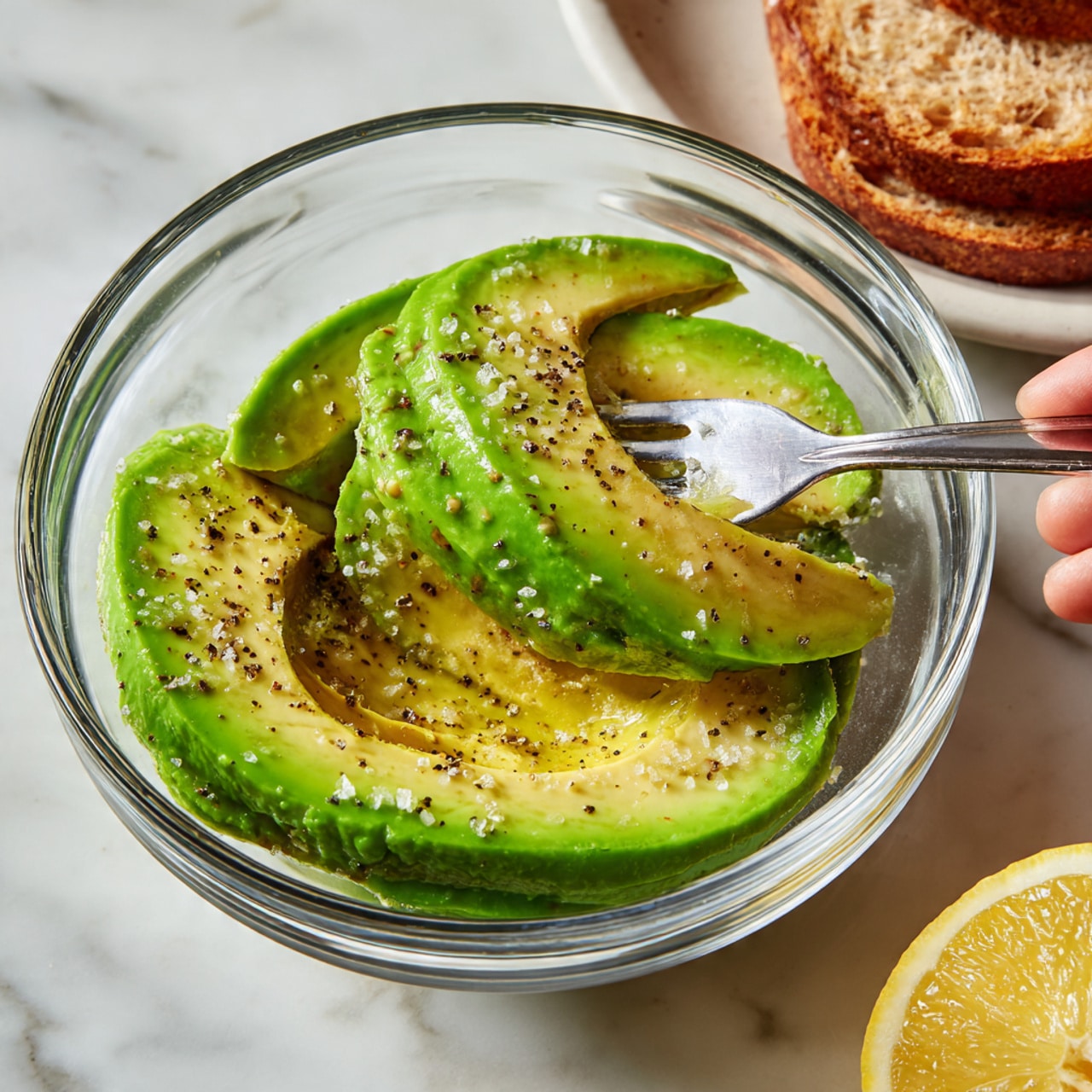 A clear glass bowl holds three slices of bright green avocado layered over each other with a smooth and creamy texture. The avocado pieces are topped with coarse black pepper and flaky sea salt crystals that add texture and contrast. The bowl is on a white marbled surface with a woman's hand holding a fork to the right, a half lemon cut with a yellow rind and juicy inside on the lower right, and some slices of toasted brown bread on a white plate in the upper right. Photo taken with an iphone --ar 4:5 --v 7