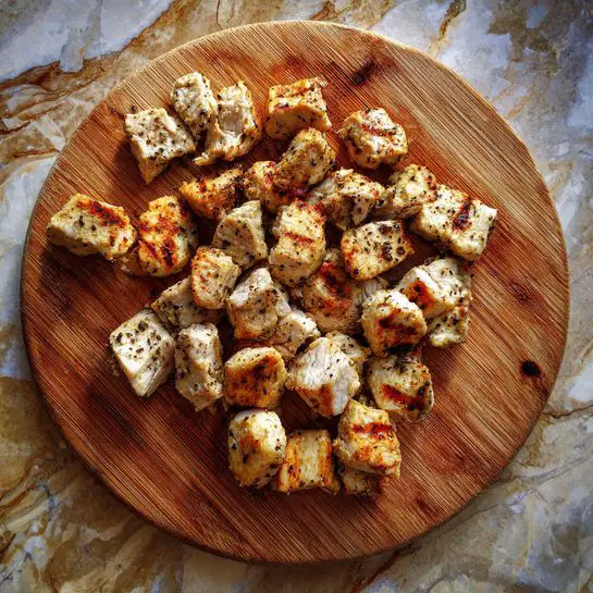 The image shows many small pieces of cooked white meat with golden brown grilled marks scattered on a round wooden cutting board. The meat pieces have a lightly textured surface with some black pepper seasoning visible. The cutting board has natural wood grain lines and knots, placed on a background with a white marbled texture. photo taken with an iphone --ar 4:5 --v 7