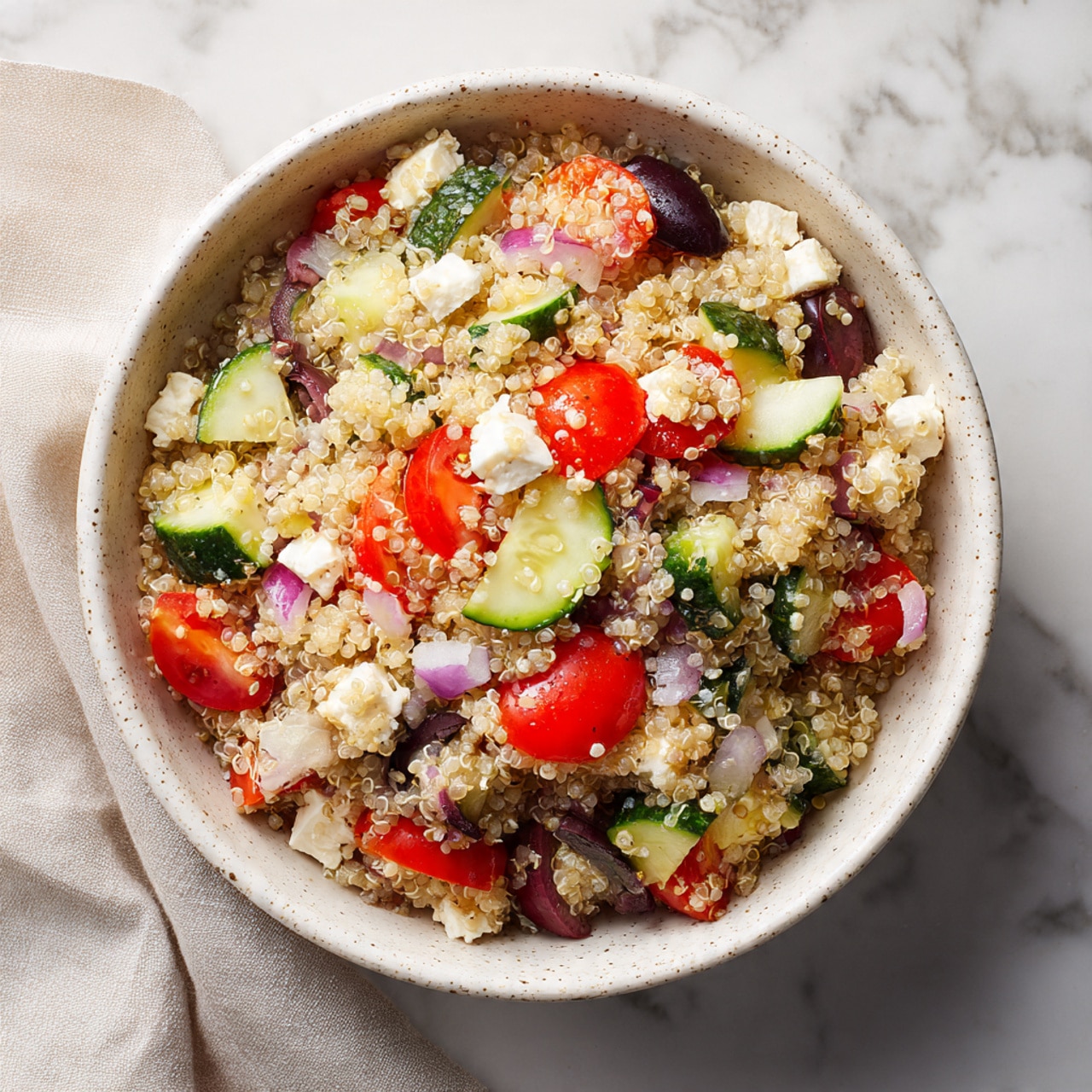A white bowl filled with a mixed quinoa salad is shown from above on a white marbled surface. The salad has multiple layers of light beige quinoa grains as the base, with scattered small cubes of white feta cheese, dark brown to black pitted olives sliced in halves, bright red cherry tomato halves, light green chopped cucumbers, small pieces of green bell pepper, and finely diced red onions spread all over. The textures range from soft quinoa to smooth and firm cheese, juicy tomatoes, and crisp cucumbers. A white cloth napkin is placed near the bowl, adding softness to the scene. Photo taken with an iphone --ar 4:5 --v 7