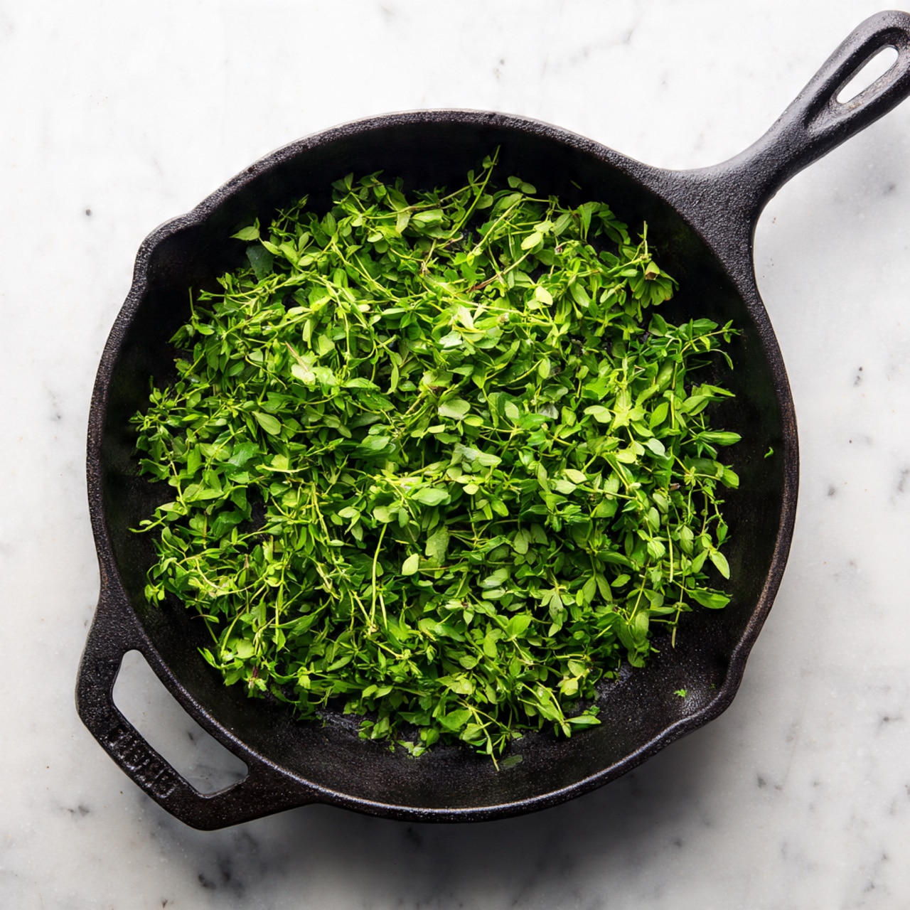 A top-down view of a black cast iron pan filled with freshly chopped green herbs spread evenly inside. The pan handle extends out to the top right corner. The pan sits on a white marbled surface that contrasts with the dark pan and bright green herbs. The herbs have a fresh, moist texture, lying flat and showing small leaves and stems in clusters. Photo taken with an iphone --ar 4:5 --v 7