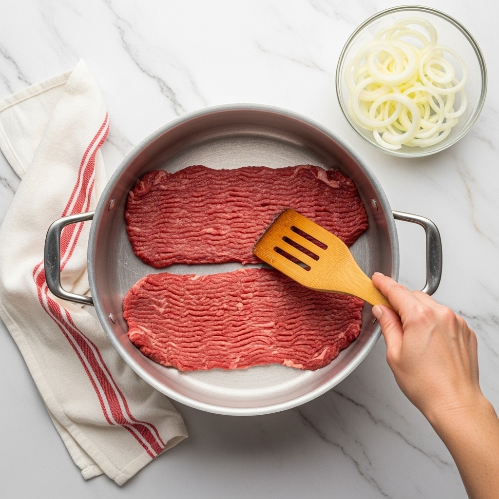 The image shows a top view of a large silver cooking pot on a white marbled surface. Inside the pot, there are two flat layers of raw red meat strips placed next to each other, with the texture showing small, fine lines from the meat fibers. A woman's hand is holding a wooden spatula with three slits, pressing down gently on the meat near the right side of the pot. On the upper right side of the image, there is a clear glass bowl filled with thin, white, circular onion slices. To the left of the pot, a white and red striped cloth is partially folded and laid on the white marbled surface. photo taken with an iphone --ar 4:5 --v 7