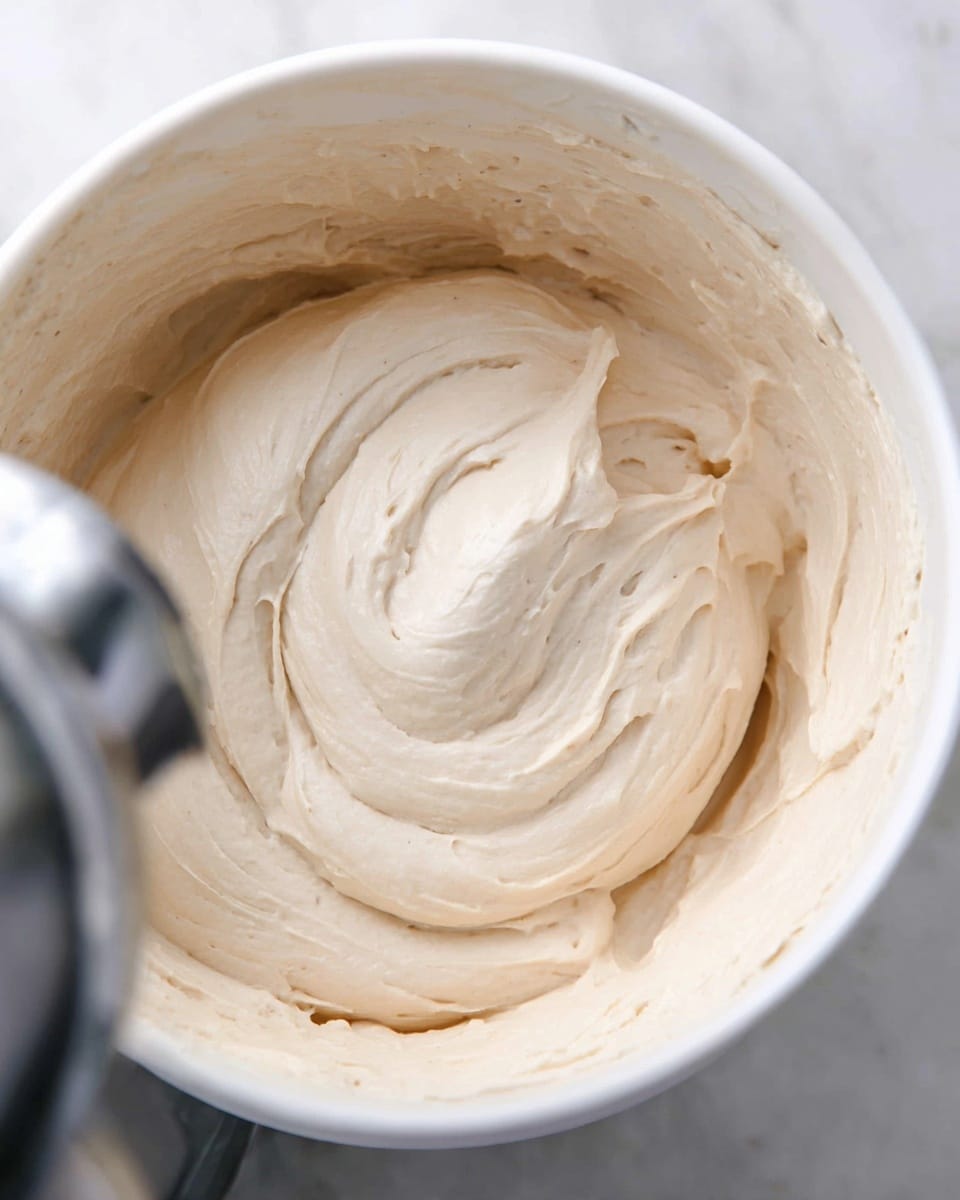 A close-up image of a thick, smooth cream with a light beige color in a white bowl. The cream has a soft, whipped texture with small peaks and swirls made by a metal mixing blade partially visible in the left side of the bowl. The overall look is creamy and soft, filling the bowl evenly. The bowl sits on a white marbled surface photo taken with an iphone --ar 4:5 --v 7