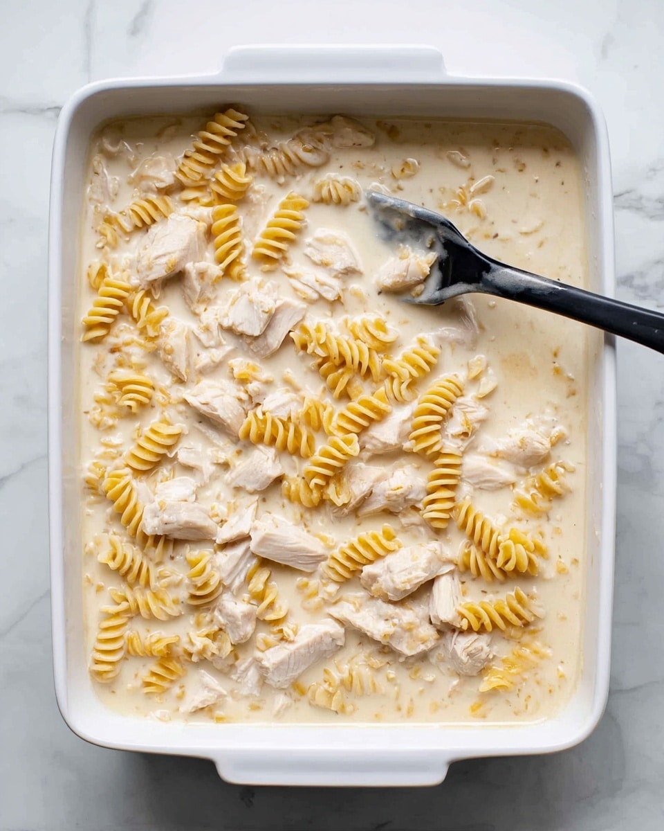 A white rectangular baking dish is filled with a creamy sauce covering two main layers: scattered pieces of light-colored cooked chicken and small spirals of dry yellow pasta submerged in the sauce. A black spatula rests inside the dish, partially covered by the sauce, positioned towards the right side. The dish sits on a surface with a white marbled texture. Photo taken with an iphone --ar 4:5 --v 7
