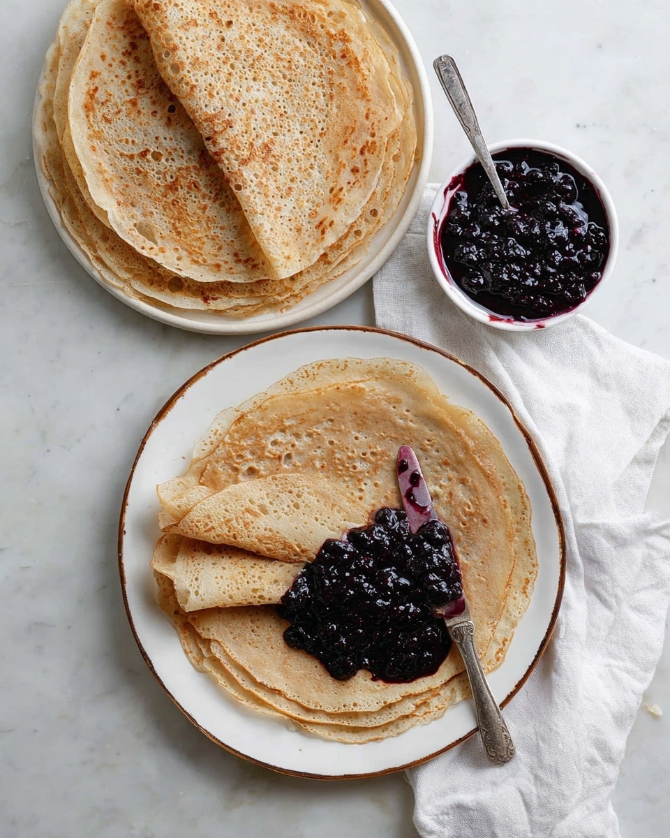 The image shows two white plates with a brown rim, each holding a stack of thin, light brown crepes with a slightly uneven texture. The plate at the bottom center of the image has a few crepes stacked, with the top crepe spread with a thick, dark purple-blue berry jam, and a silver butter knife resting on top with some jam on its blade. The second plate is placed at the top and holds a similar stack of plain crepes. A small white bowl filled with the same dark berry jam is placed at the top right. The items are arranged on a white marbled surface with a white cloth napkin beside the plates. Photo taken with an iphone --ar 4:5 --v 7