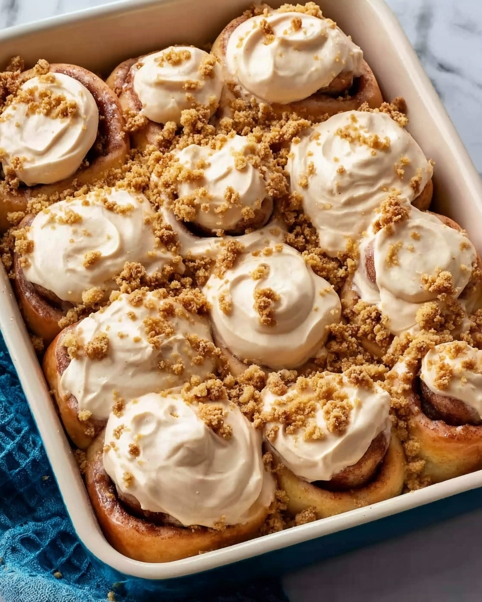 The image shows a white rectangular baking pan filled with nine cinnamon rolls. Each cinnamon roll has a thick layer of creamy light brown frosting spread on top, with some areas showing smooth texture and slight swirls. Crumbly streusel bits of light golden brown color are sprinkled around and slightly on top of the frosting. The cinnamon rolls are soft and golden brown, tightly rolled with visible cinnamon swirls inside. The pan is placed on a white marbled surface with a blue cloth partially visible beside it. photo taken with an iphone --ar 4:5 --v 7