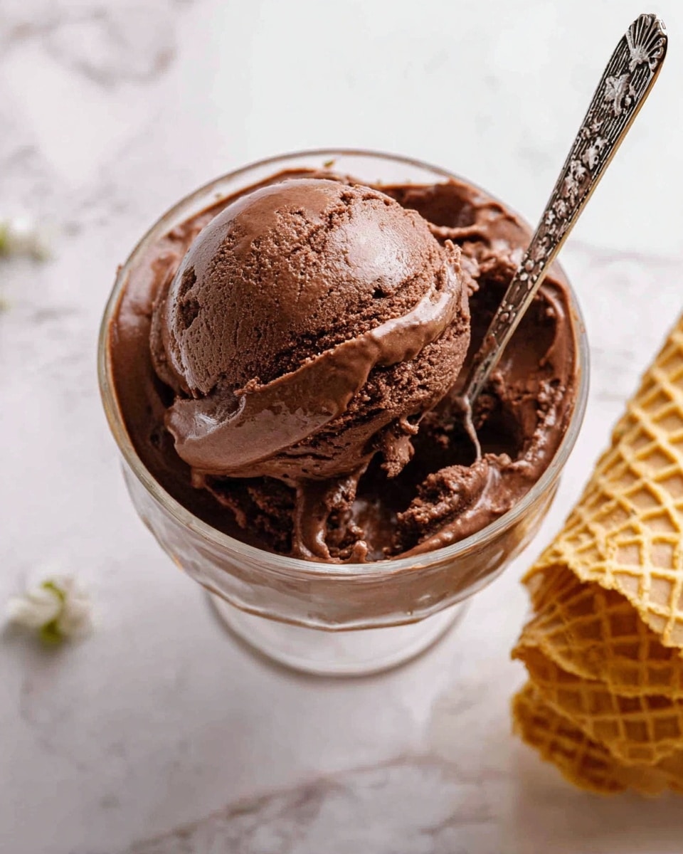 A close-up view of a large scoop of rich, dark brown chocolate ice cream with a smooth and slightly creamy texture, served in a clear glass bowl. A silver spoon with intricate designs is inserted into the top of the ice cream, ready for eating. To the right side of the image, there is a stack of golden waffle cones with a crisp texture. The bowl is placed on a surface with a white marbled pattern, giving a clean and simple background. Photo taken with an iphone --ar 4:5 --v 7