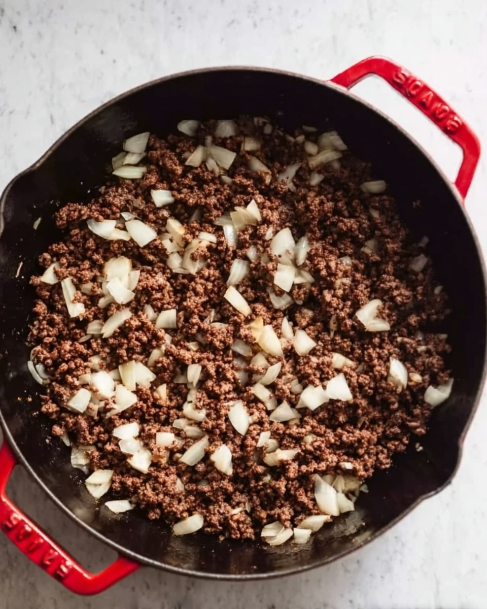A close-up view of a black cast iron pan filled with browned ground meat mixed with chunks of cooked white onions. The pan has red handles and sits on a white marbled surface. The ground meat is evenly spread across the pan, with the onions scattered throughout, showing a mix of brown and white colors and a coarse texture. The background is soft and clean, emphasizing the cooked ingredients inside the pan. Photo taken with an iphone --ar 4:5 --v 7