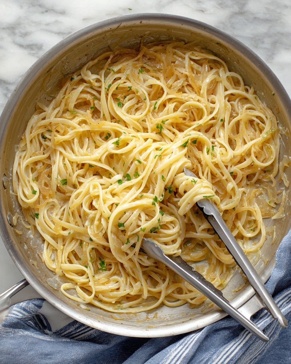 A large silver pan filled with creamy pasta mixed with soft cooked onion strands. The pasta is light yellow with a smooth and slightly shiny texture, tangled inside the pan, and the onions are a translucent golden color, spread evenly throughout the noodles. Some finely chopped green herbs are scattered subtly on top, adding a hint of color contrast. A pair of silver tongs rests inside the pan, partially buried in the pasta. The pan sits on a white marbled surface with a folded blue and white striped cloth partially visible underneath it. Photo taken with an iphone --ar 4:5 --v 7