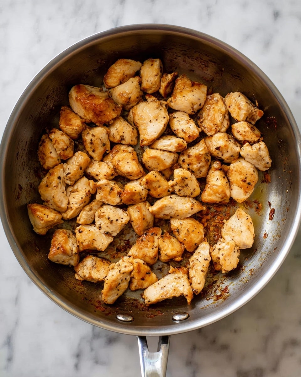 The image shows a single layer of small, golden brown cooked chicken pieces evenly spread inside a round stainless steel pan with two handles, sitting on a white marbled surface. The chicken pieces have a slightly crispy texture with some black pepper scattered over them. The pan has a shiny interior with some cooked juices and small browned bits visible on the surface around the chicken. Photo taken with an iphone --ar 4:5 --v 7