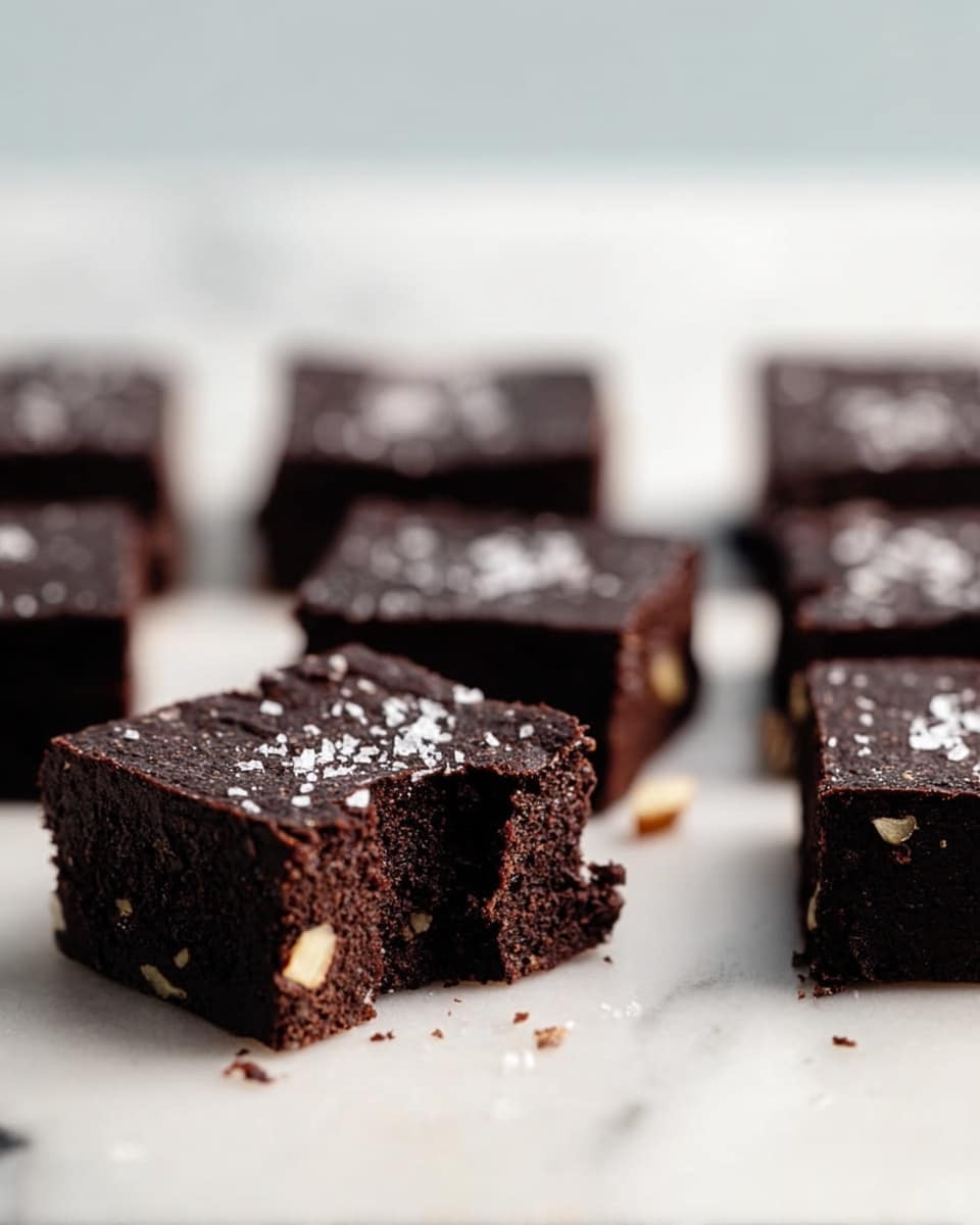 The image shows several small square dark chocolate brownies placed closely on a white marbled surface. The brownies have a rich dark brown color with a slightly rough texture on top, sprinkled lightly with white salt flakes. One brownie in the front has a bite taken out, showing a dense and moist inside with small light brown nut pieces scattered throughout. The brownies are arranged in a way that some are in the background, slightly blurred. Photo taken with an iphone --ar 4:5 --v 7