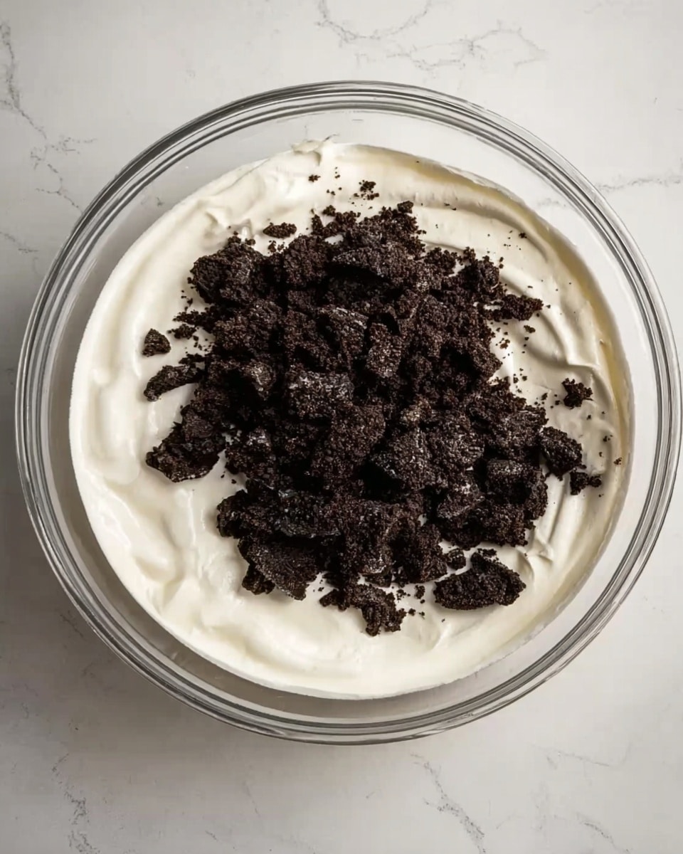 A clear glass bowl filled with a thick, smooth white cream base that covers most of the bowl, with dark brown crushed cookie pieces piled in the center on top of the cream. The textures show the creamy layer as soft and fluffy, while the cookie pieces look crunchy and uneven in size. The bowl is placed on a white marbled surface. photo taken with an iphone --ar 4:5 --v 7