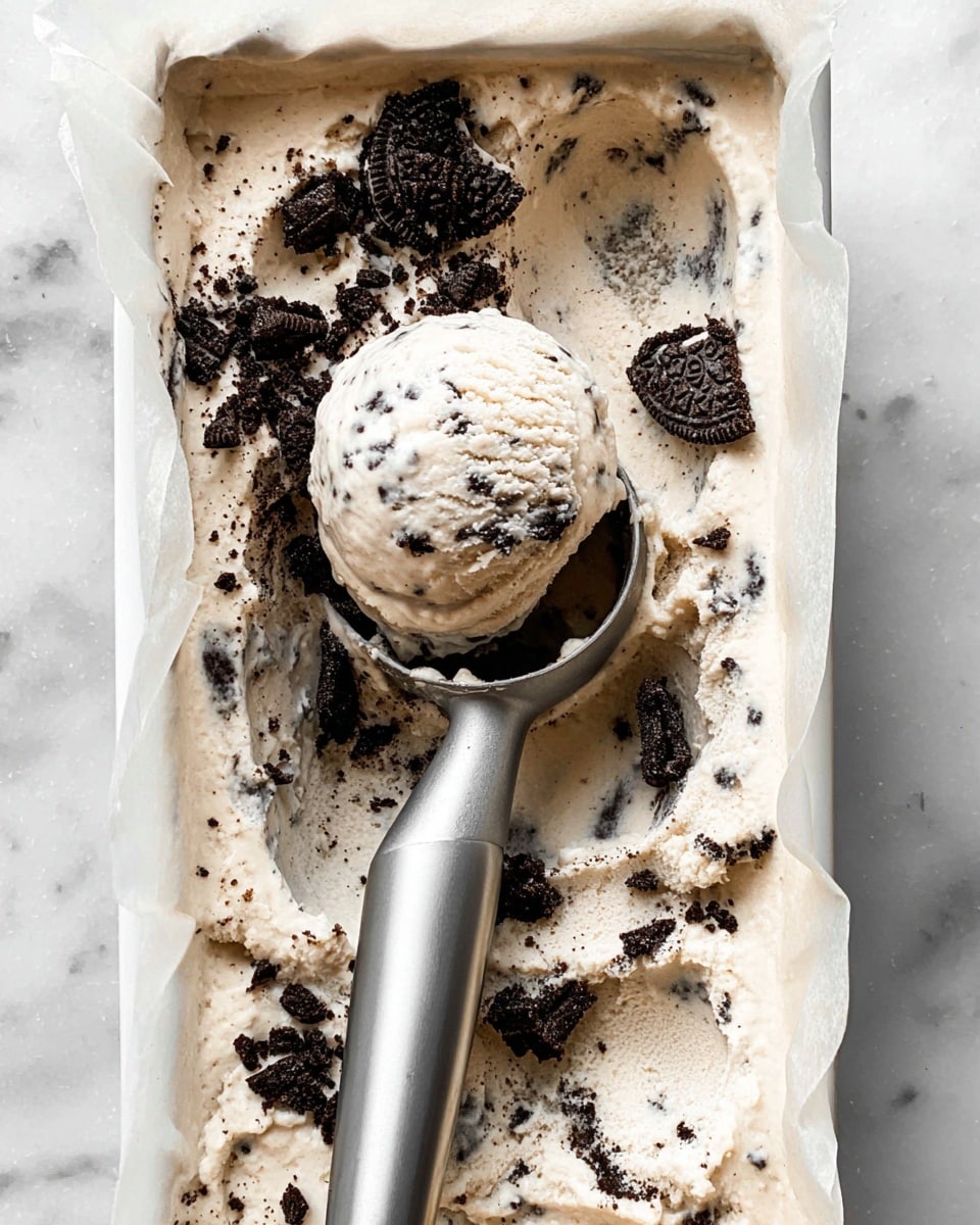 A white rectangular container lined with parchment paper holds creamy cookies and cream ice cream, filled with visible chunks of black cookie pieces throughout. A silver ice cream scoop in the middle lifts a rounded scoop of the pale off-white ice cream speckled with cookie bits. The ice cream has a soft, smooth texture with crushed dark cookies scattered on one side. The background is a white marbled surface. photo taken with an iphone --ar 4:5 --v 7
