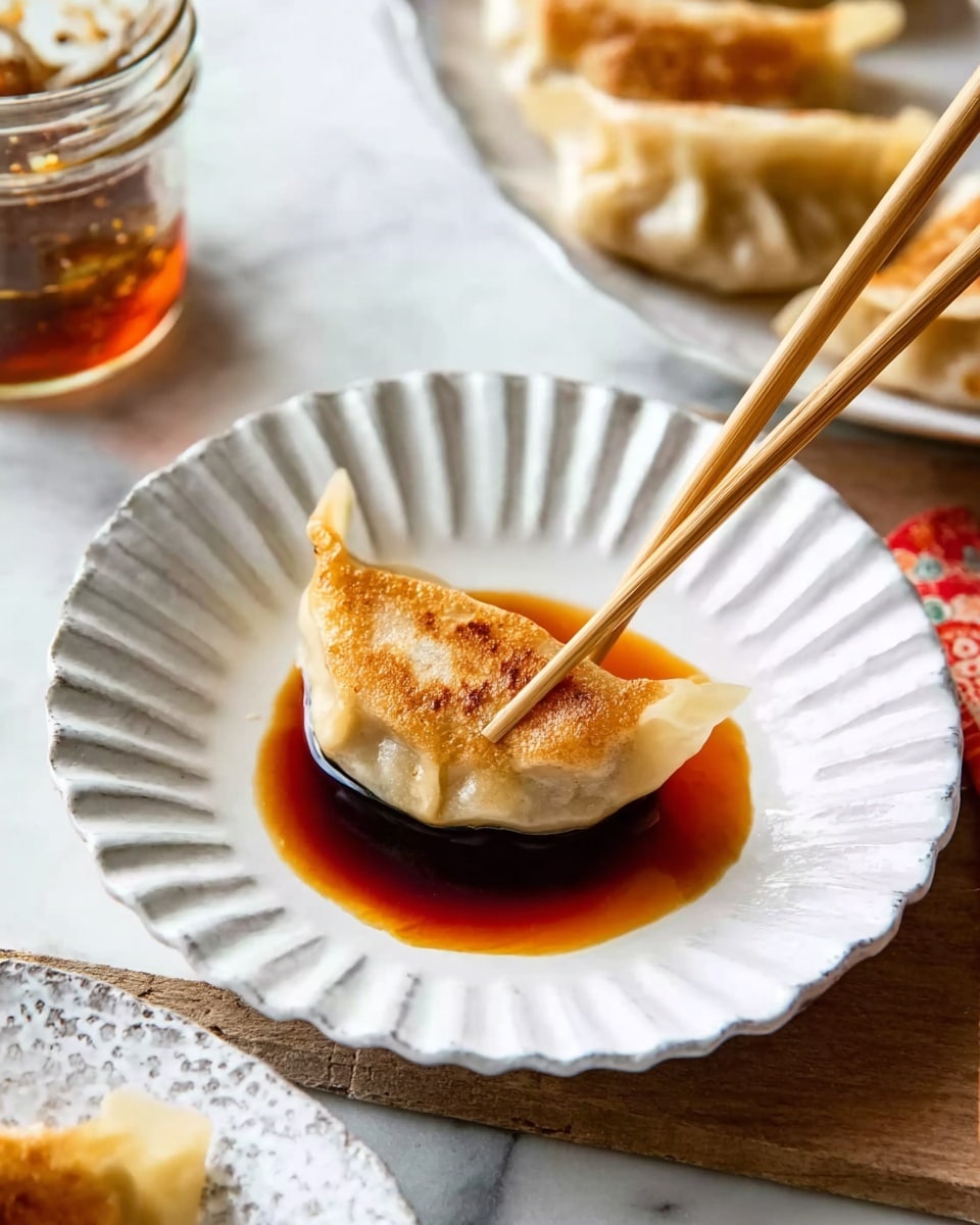 A white fluted plate holds a single golden-brown dumpling resting in a shallow pool of dark soy dipping sauce that spreads across the plate’s base. The dumpling’s crispy bottom contrasts with its soft, slightly translucent upper dough. A pair of wooden chopsticks, held by a woman’s hand, gently pinches the dumpling from above, angled diagonally into the sauce. The plate sits on a white marbled surface with a glimpse of another white fluted plate with more dumplings off to the side, and a partially visible glass jar of sauce nearby. photo taken with an iphone --ar 4:5 --v 7