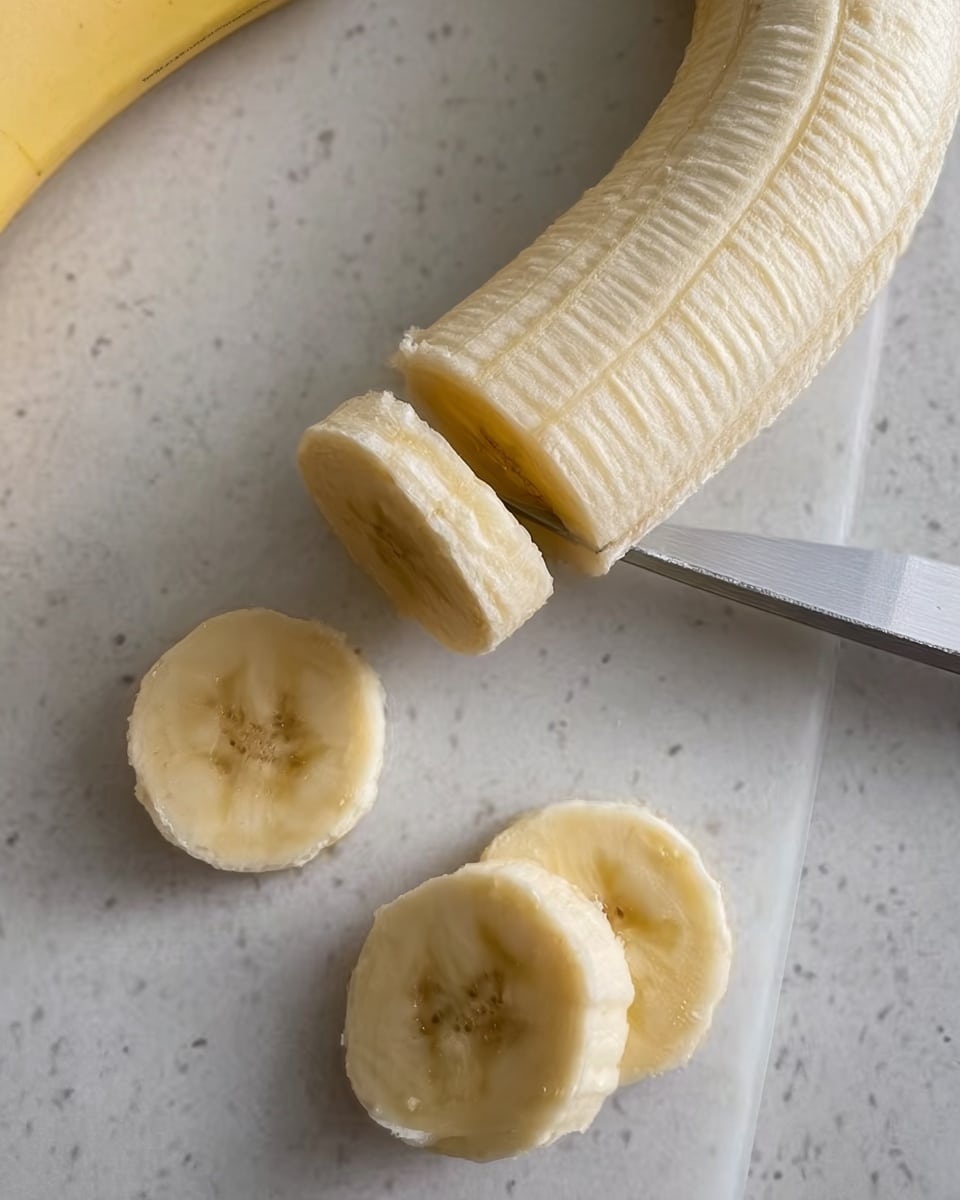 The image shows a peeled banana being sliced on a white marbled surface. One whole banana is placed horizontally, and a knife is slicing a piece from the banana closest to the bottom of the image. Two round slices of banana with a soft yellow inside and a smooth texture are already cut and placed near the bottom left corner. The banana has a light creamy yellow color with a soft texture, and the knife blade is metallic and sharp. Photo taken with an iphone --ar 4:5 --v 7