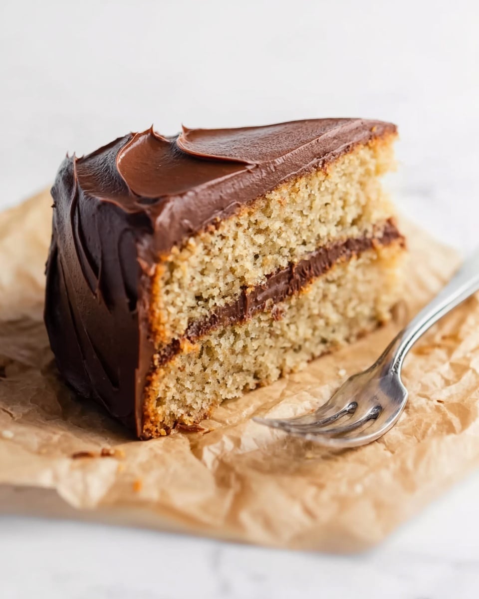 A slice of two-layer cake with light brown textured sponge layers and a smooth, thick dark chocolate frosting covering the top and sides, showing soft swirls on the outside. The cake slice rests on crumpled light brown parchment paper, accompanied by a silver fork on the right. The whole scene is set on a white marbled surface. photo taken with an iphone --ar 4:5 --v 7