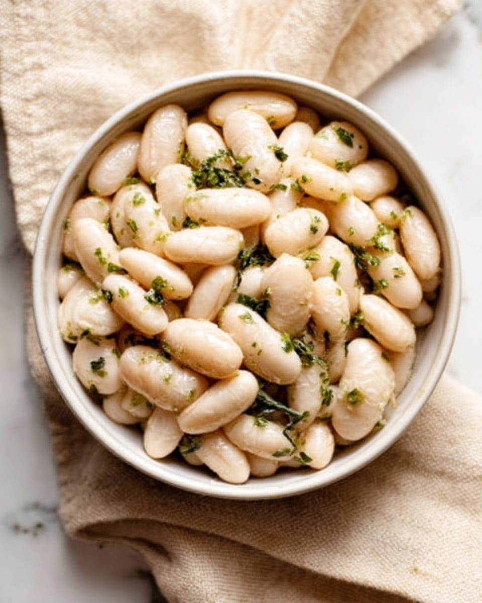 The image shows a white bowl filled with large white beans mixed with small green herb pieces. The beans have a soft, slightly shiny texture and are closely packed inside the bowl, which sits on a beige cloth napkin. The background surface is a white marbled texture. The scene looks bright and clean. photo taken with an iphone --ar 4:5 --v 7