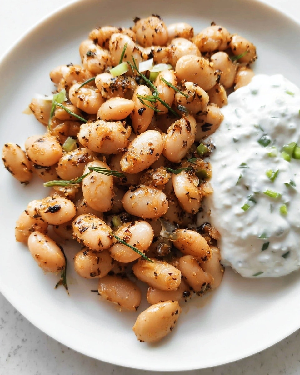 The image shows a pile of cooked white beans seasoned with visible dark herbs and small pieces of green onions, all with a light brown toasted texture, spread on a white plate. On the right side of the plate, there is a dollop of creamy white sauce with small green herb pieces mixed in. The surface around the plate has a white marbled texture. Photo taken with an iphone --ar 4:5 --v 7