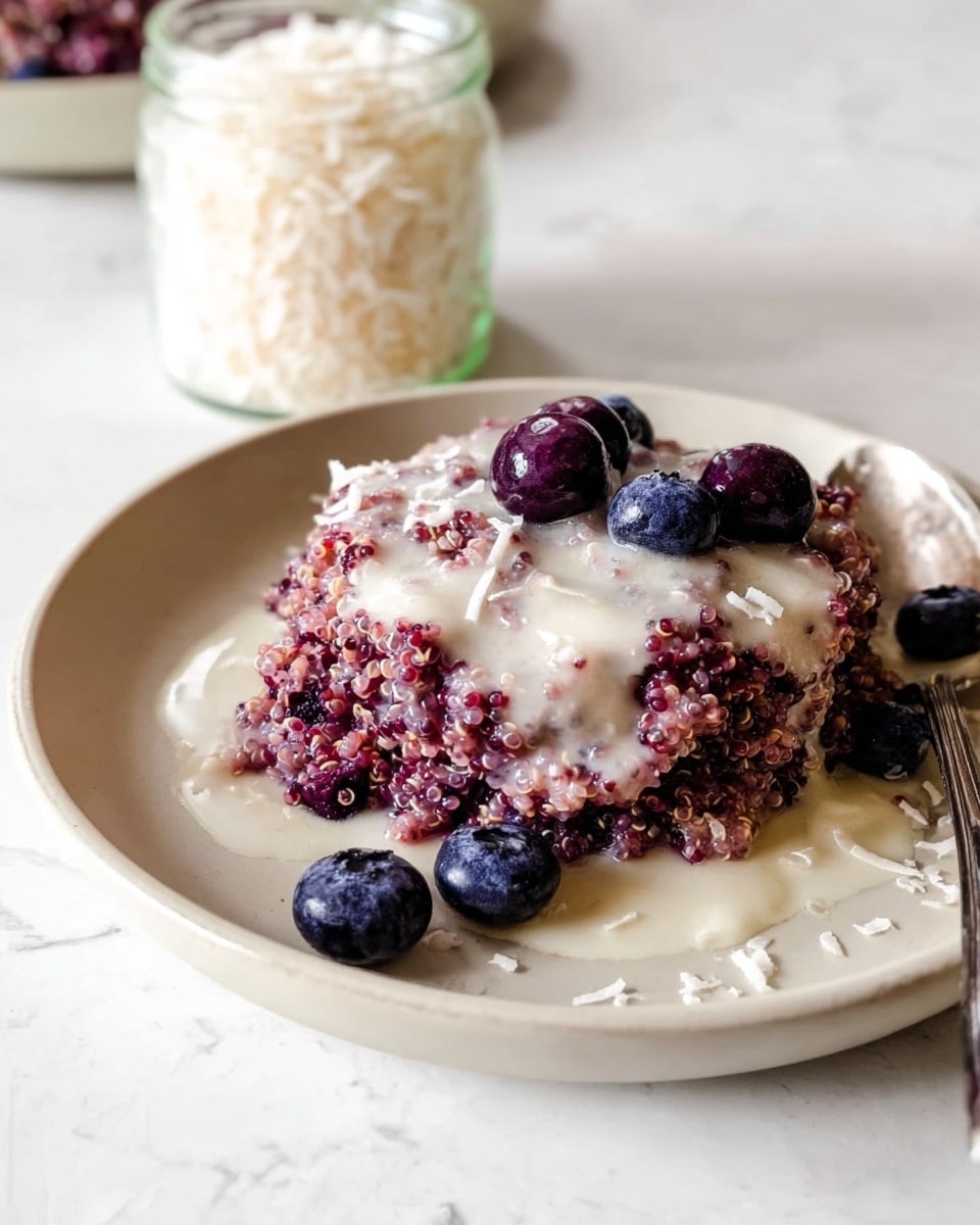 A round white plate holds a stack of purple quinoa with mixed berries, creating a textured layer with a mix of deep purple and light pink hues. On top, a layer of creamy white sauce is drizzled, softly blending into the quinoa beneath it. Several plump blueberries and darker berries are scattered on the creamy surface, adding small pops of deep blue and dark red. The plate sits on a white marbled surface. In the top left, a glass jar filled with shredded coconut is visible. A silver spoon peeks from the right side of the plate. Photo taken with an iphone --ar 4:5 --v 7