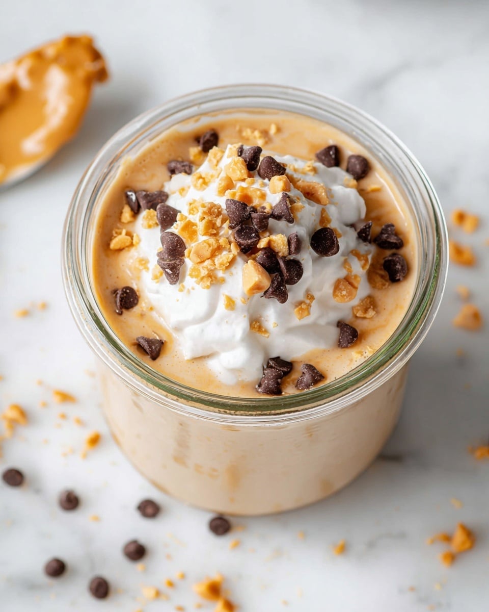 A glass jar filled with a creamy light brown base layer topped with a scoop of white whipped cream, sprinkled with small dark brown chocolate chips and crushed light golden peanut pieces. The jar is placed on a white marbled surface, with some scattered crumbs nearby. Photo taken with an iphone --ar 4:5 --v 7