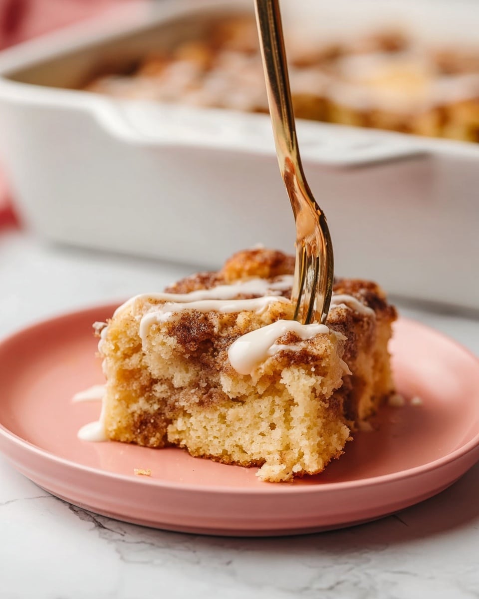 A piece of crumbly cake sits on a pale pink plate with a fork pressed into it from the top. The cake has two visible layers: a light tan, soft, crumbly base with darker golden-brown areas suggesting cinnamon or spice, and a thin drizzle of white icing on top. The background shows a white baking dish with more cake inside, resting on a white marbled surface. Photo taken with an iphone --ar 4:5 --v 7