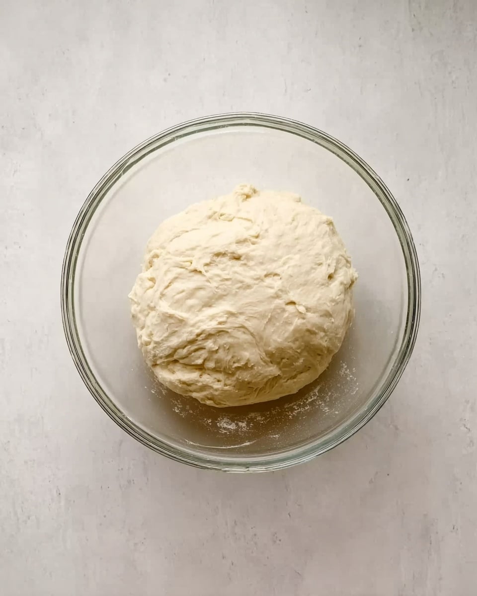A clear glass bowl sits on a white marbled surface, holding a ball of smooth, pale dough with slight uneven textures on its surface. The dough fills the center of the bowl, showing a soft and slightly shiny appearance. Photo taken with an iphone --ar 4:5 --v 7