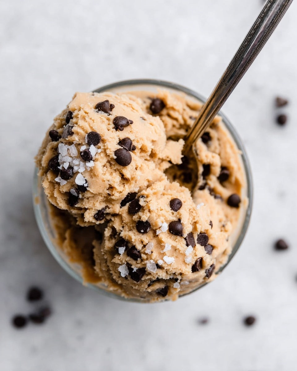 The image shows a clear glass bowl filled with two generous scoops of light brown cookie dough mixed with small, dark chocolate chips evenly spread throughout. The cookie dough is topped with a sprinkle of coarse sea salt crystals, adding a touch of texture on the surface. A metallic spoon is inserted into the cookie dough, positioned diagonally from the top right to the center. The bowl sits on a white marbled surface with a few scattered small chocolate chips around it, creating a clean and simple setting. photo taken with an iphone --ar 4:5 --v 7