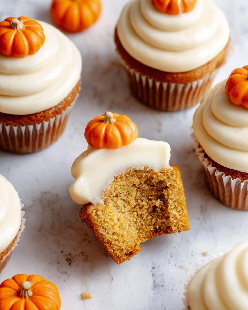 The image shows several cupcakes on a white marbled surface, each with two layers: the bottom layer is golden brown with a soft, moist texture, and the top layer is smooth creamy white frosting swirled in a spiral. Each cupcake is topped with a small orange pumpkin decoration that is round with subtle ridges and a tiny dark stem. One cupcake is shown with a bite taken out, revealing the inside texture of the cake. The bright orange pumpkins are scattered around the cupcakes, adding a pop of color to the scene. photo taken with an iphone --ar 4:5 --v 7