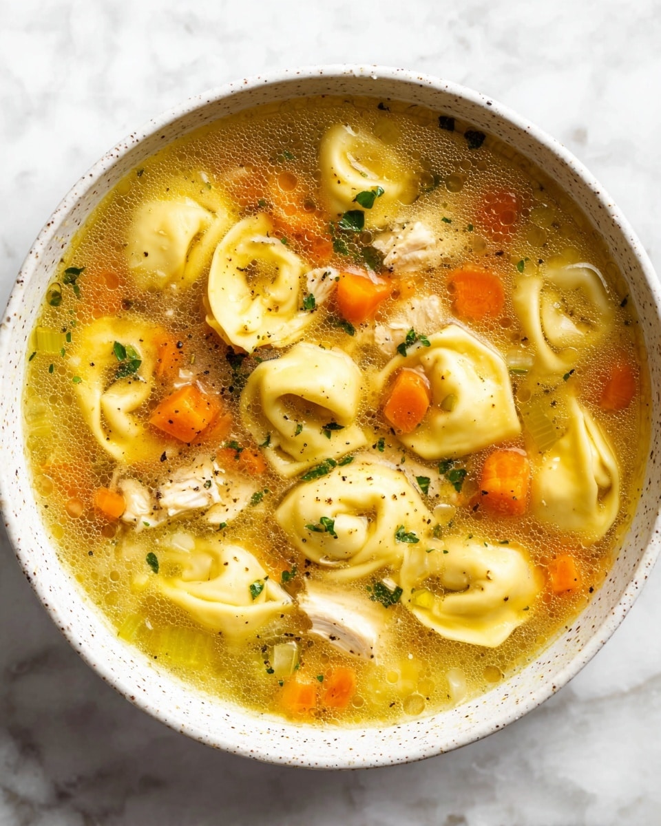 A white speckled bowl filled with clear golden broth layered with soft yellow tortellini scattered evenly throughout. There are small diced orange carrots and light green celery pieces in the soup, mixed with shredded white chicken. Tiny bits of fresh green herbs float on top, adding color contrast. The broth shows slight bubbles and a sheen of oil on the surface. The bowl is set on a white marbled texture surface. photo taken with an iphone --ar 4:5 --v 7