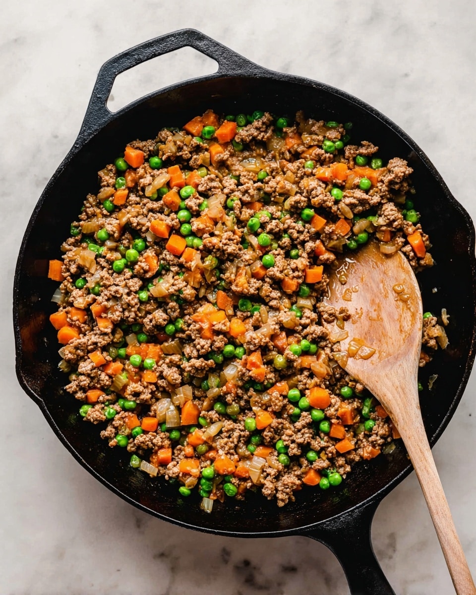 Inside a black cast iron skillet, there is a mix of cooked ground meat, green peas, and orange carrot cubes scattered evenly throughout. The mixture has a slightly shiny, saucy texture, and bits of tender, cooked onions are visible, adding a light brown color with soft texture. A wooden spoon rests on the right side of the skillet, partly covering the food, with the handle extending out of the skillet. The skillet is placed on a white marbled surface. photo taken with an iphone --ar 4:5 --v 7