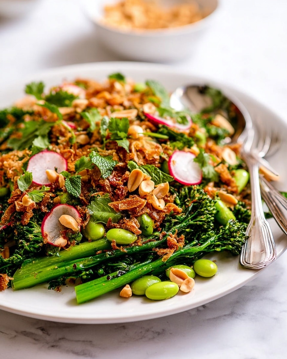 A close-up image of a white plate filled with a colorful salad made of several layers. The bottom layer is bright green broccolini stalks, which look fresh and crisp. On top of the broccolini, there is a mix of whole and chopped light green edamame beans and thin slices of red radish with white centers. The top layer consists of golden brown crispy fried shallots or onions, adding a crunchy texture, scattered with broken pieces of light brown peanuts. Small green mint leaves and fresh light green cilantro bits are sprinkled throughout, adding liveliness and freshness. The plate is placed on a white marbled surface, with a silver fork and spoon resting on the right side. The background shows a blurred white bowl with more peanuts. Photo taken with an iphone --ar 4:5 --v 7