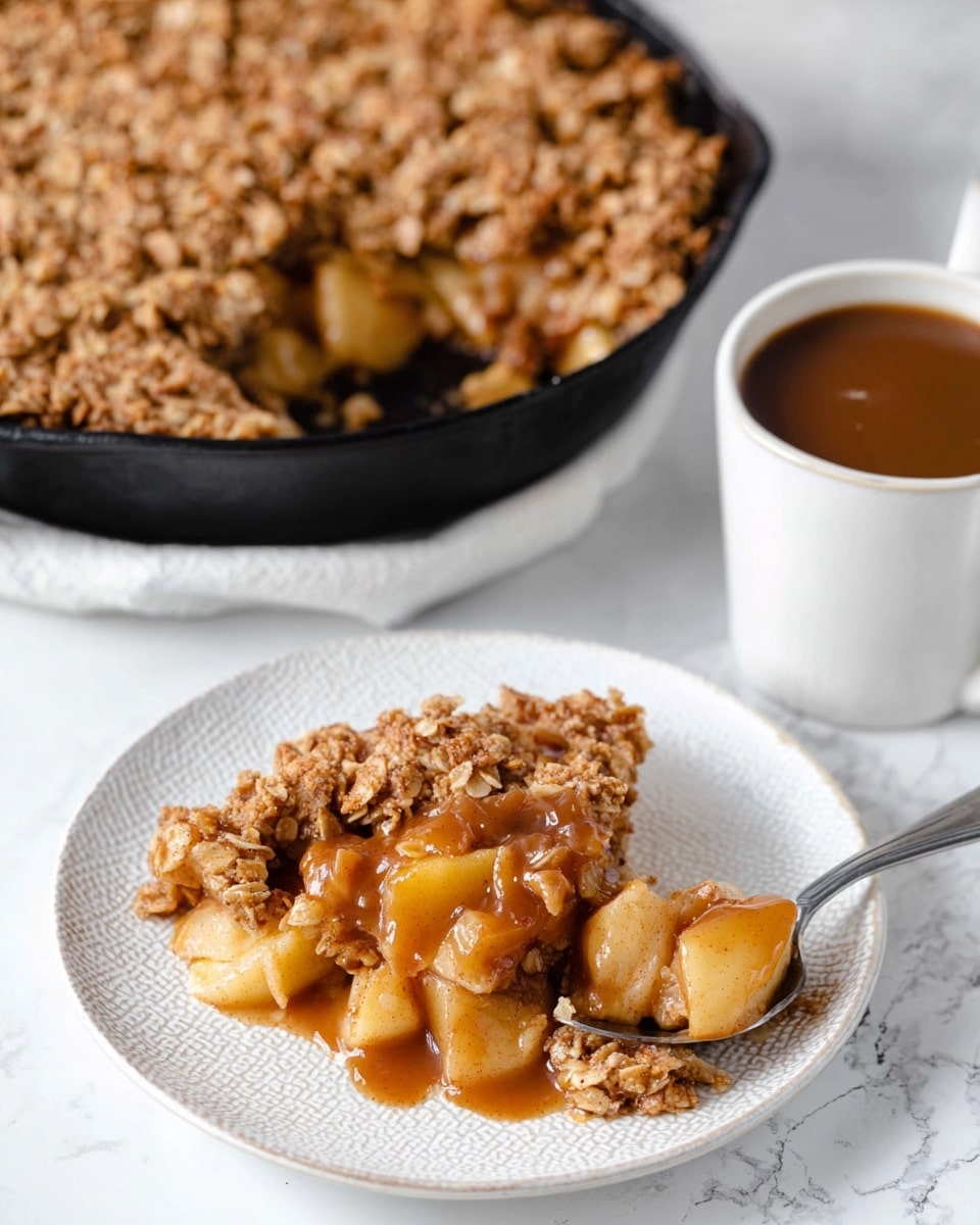 The image shows a slice of apple crisp dessert on a white plate with a textured rim. The dessert has two layers: the bottom layer is made of chunky, soft apple pieces mixed with a cinnamon syrup that is shiny and brown, and the top layer is a golden brown oat crumble that looks crunchy and thick. Next to the plate, there is a silver spoon holding some of the apple crisp. In the background, a black cast iron skillet holds the rest of the apple crisp, with the oat crumble and cinnamon syrup visible on top. To the right, there is a white mug filled with brown sauce, all placed on a white marbled textured surface. Photo taken with an iphone --ar 4:5 --v 7
